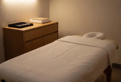 A clean and minimalist empty room prepared for acupuncture therapy, featuring a treatment bed with white linens, wooden accents, and soft lighting for relaxation.