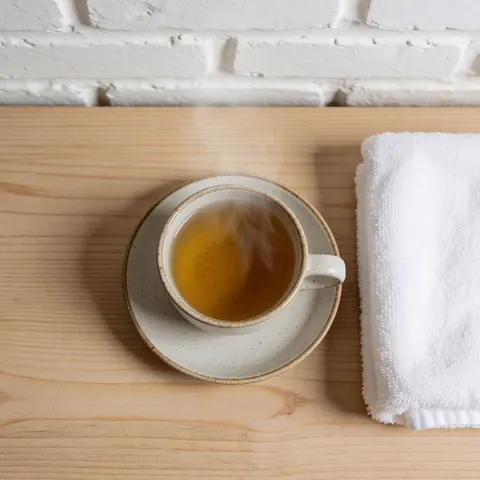 A ceramic cup of steaming chamomile tea sits on a wooden table with a white towel nearby, set against a white brick wall for a calm and relaxing atmosphere.