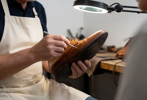 A shoemaker in a white apron repairs a brown leather shoe under magnifying light in a workshop