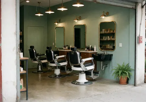 Barbershop interior featuring vintage chairs, large mirrors, green walls, a corner plant, and a sink area with chrome and wood details