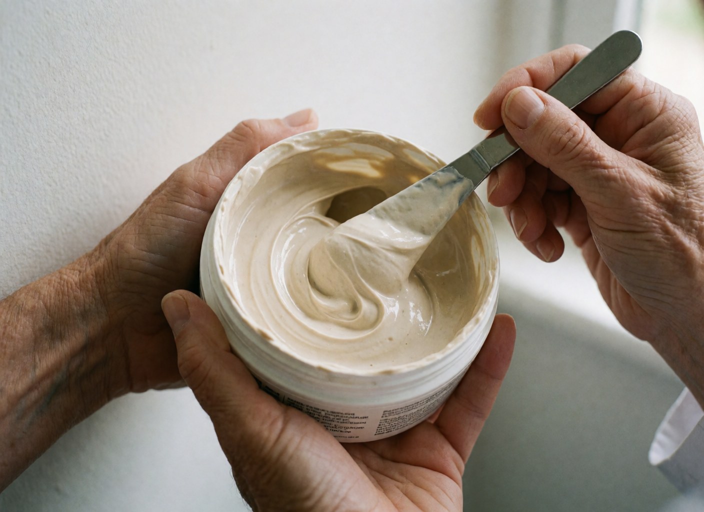 Macro close-up of beige hair mask in white jar being applied with metal spatula held by adult hands against white wall