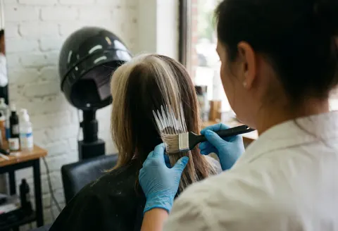 A hair stylist wearing blue gloves uses a brush to apply balayage dye to a client's hair in a salon with white brick walls and natural light