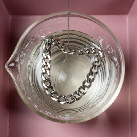 A silver chain being electroplated in a glass beaker with chemical solution on a pink background