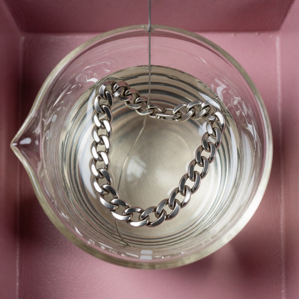 A silver chain being electroplated in a glass beaker with chemical solution on a pink background