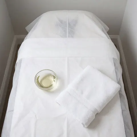 Close-up of a clean massage table covered with a white towel, featuring a small cup of oil and a folded towel, ready for a professional treatment session.