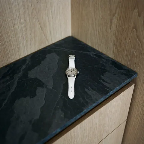 Close-up of an elegant silver dial wristwatch with a white strap resting on a dark surface next to light wood, symbolizing jewelry repair and luxury timepieces.