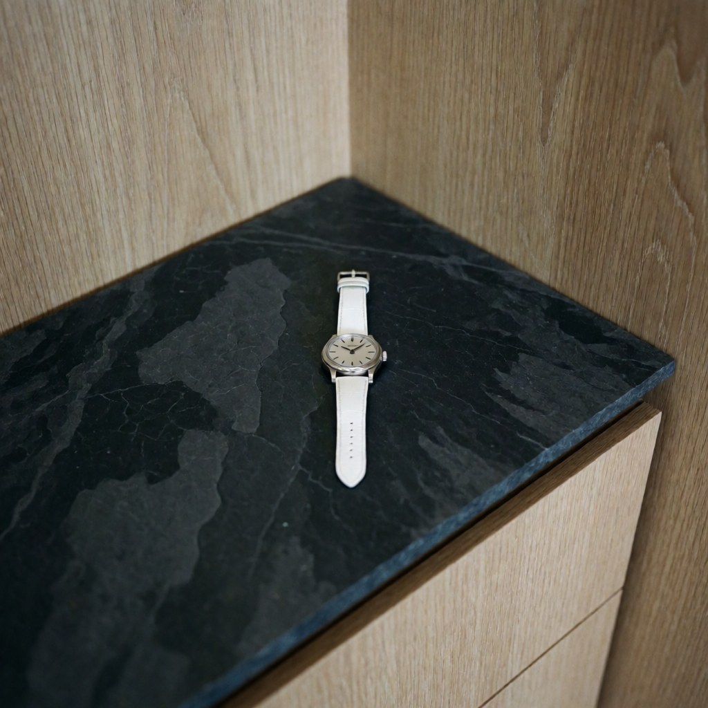 Close-up of an elegant silver dial wristwatch with a white strap resting on a dark surface next to light wood, symbolizing jewelry repair and luxury timepieces.