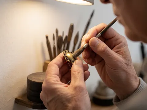 Close-up of a jeweler's hands applying polish to an antique bronze brooch using a small brush, with blurred tools in the background.