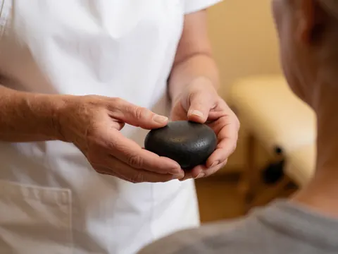 A physiotherapist with clean hands and short nails holds a warm basalt stone, preparing to apply a therapeutic shoulder massage in a relaxing treatment room.