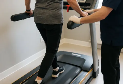 A physiotherapist wearing gloves adjusts the posture of an older patient on a treadmill in a clean, professional rehabilitation clinic.