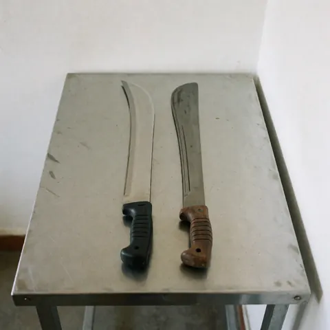 A collection of agricultural and field tools such as a machete, cane knife, and straight blade arranged on a metal table against a white backdrop for repair or product display