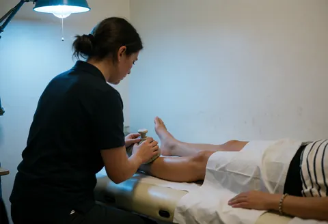 A physiotherapist applies cold gel to a patient's ankle while they rest on a treatment table in a clinical setting with blue lighting.