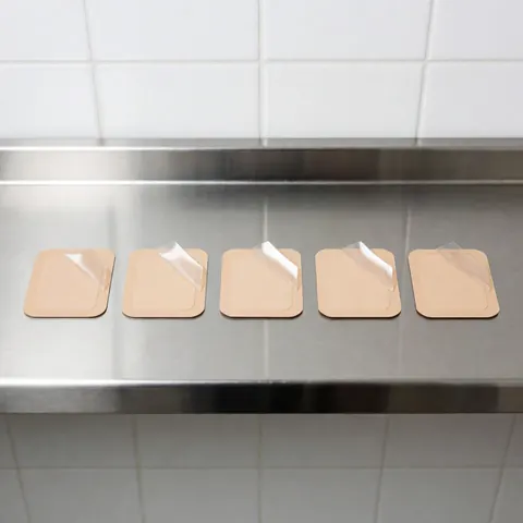 Five beige transdermal patches with partially removed clear film arranged in a row on a shiny stainless steel surface against white tiled walls, creating a clean sterile scene for osteopathy.