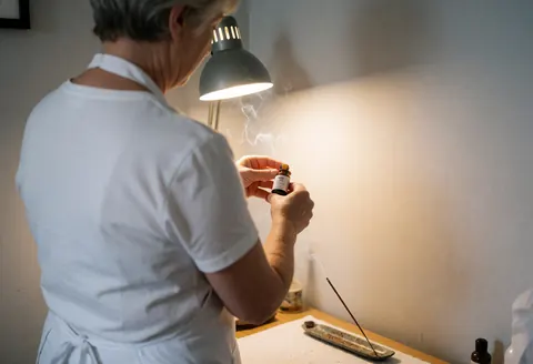 A reiki practitioner prepares lavender essential oil in a calm workspace with burning incense and warm lighting.