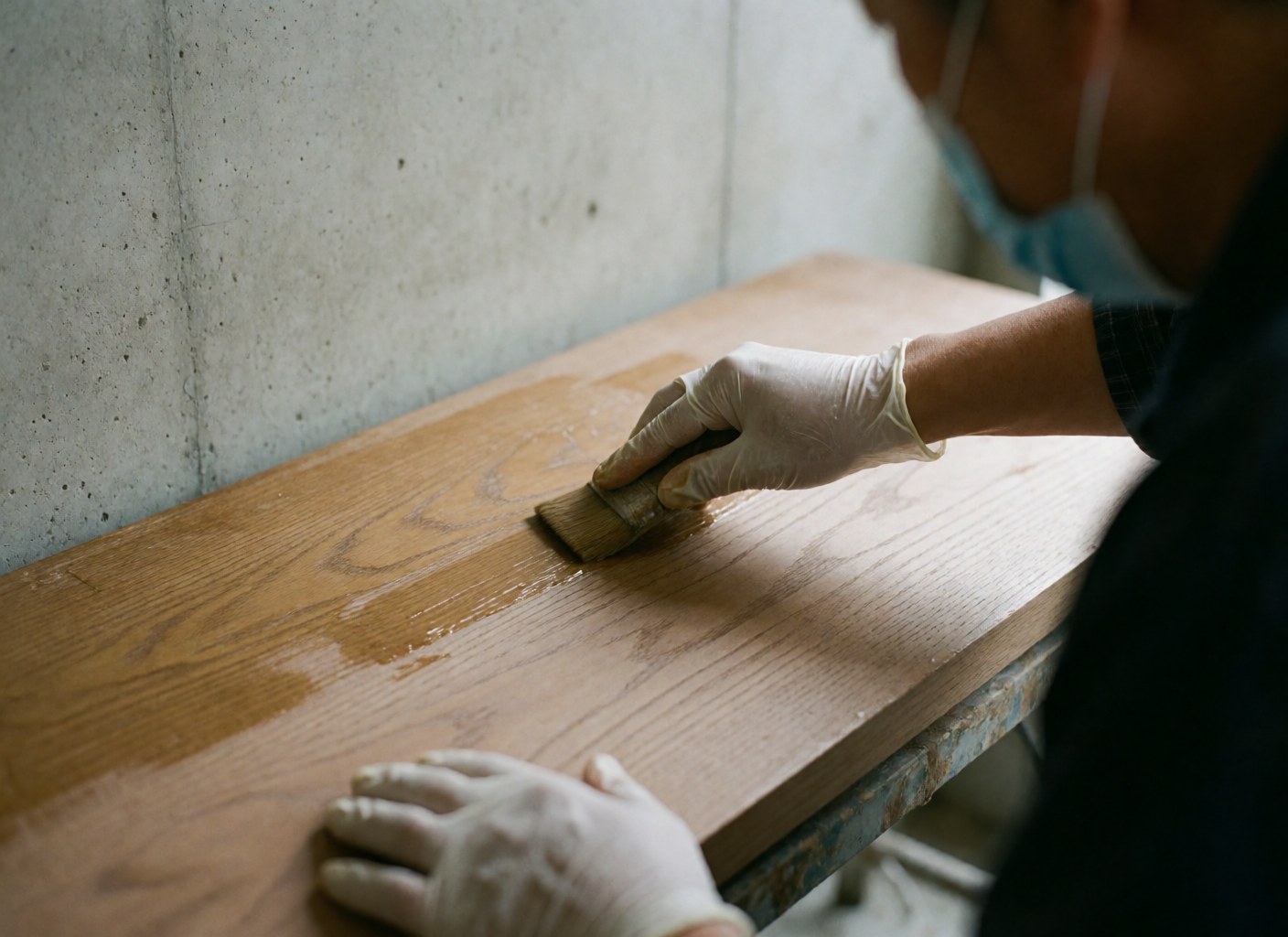 Hand wearing a glove applying matte wax with a brush on polished wood surface, concrete wall background, furniture repair scene