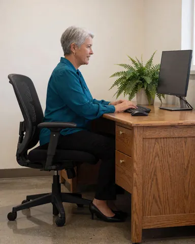 A woman sits at a wooden desk in an office, working on a computer with a plant nearby, using a keyboard and monitor, seated on an office chair.