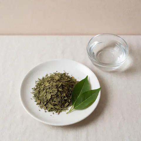 White ceramic plate holding fresh and dried coca leaves alongside a glass of water, set on light linen fabric against a soft beige background for holistic therapy products.