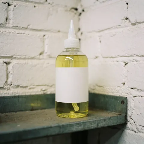 Clear plastic bottle with white dropper cap containing yellow sharpening oil, placed on a metal rack against a white brick wall for tool maintenance.