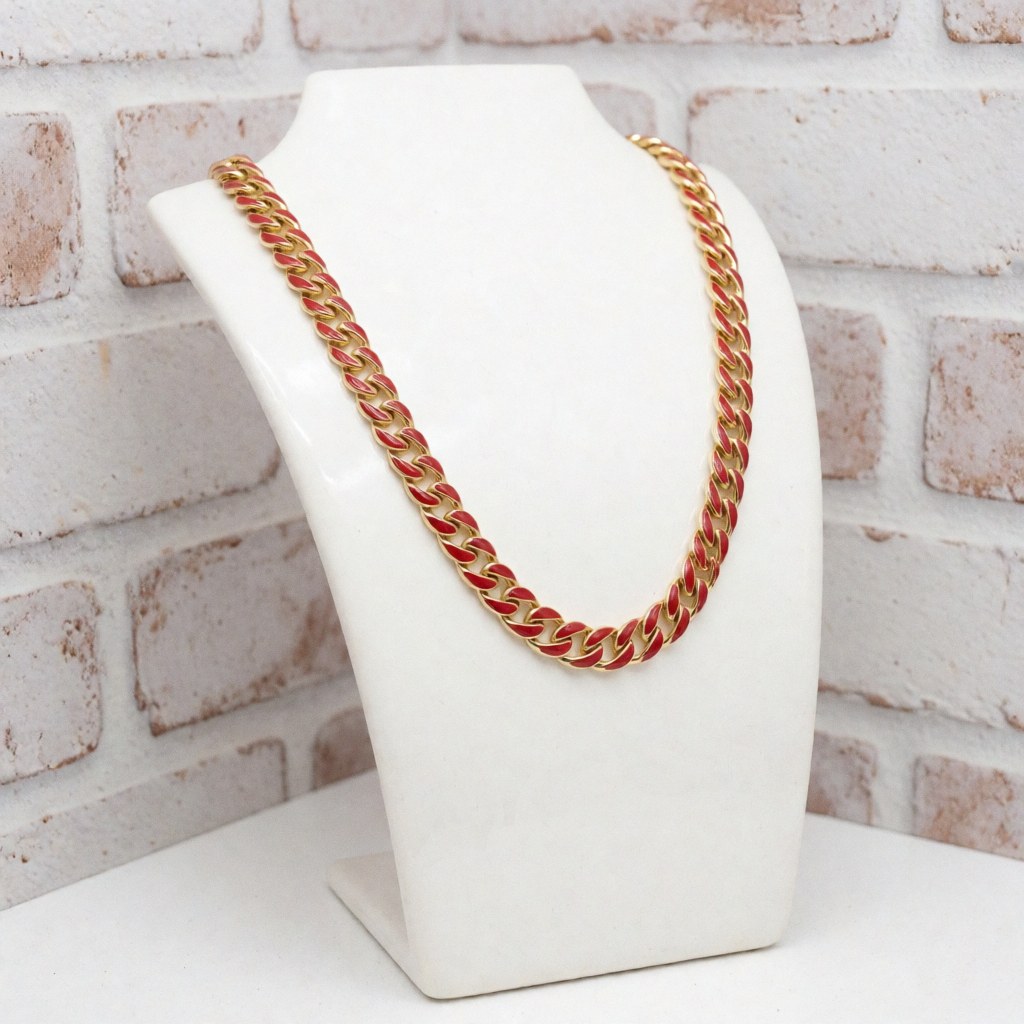 Gold necklace featuring bright red enamel displayed on a white stand against a painted brick wall, suitable for jewelry store product catalogs.