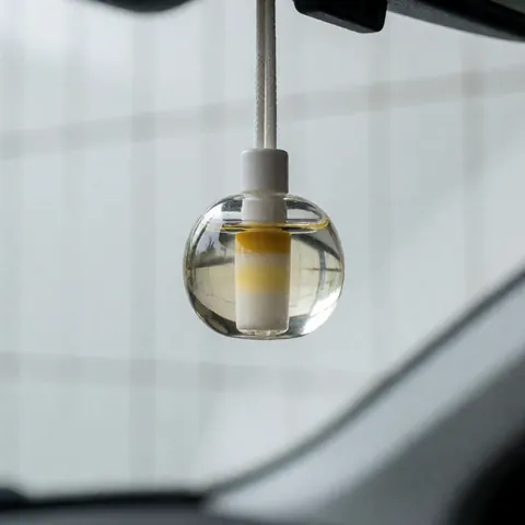 A close-up view of a modern, transparent car air freshener hanging from the rearview mirror, allowing natural light to pass through its clean design.