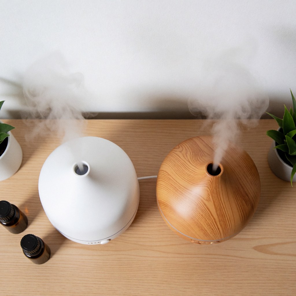 Two aromatherapy diffusers, one white and one wood, releasing soft vapor on a light wooden table with essential oil bottles and small green plants