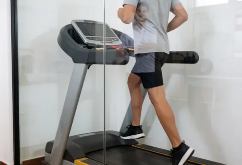 Athlete exercising on a treadmill in a therapy room, with the therapist's reflection visible in the glass.