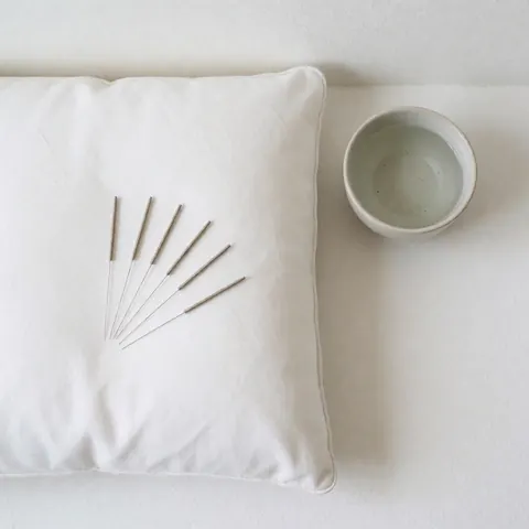 Close-up view of acupuncture needles, a small bowl of water, and meditation cushions arranged neatly on a clean white surface for therapy and wellness