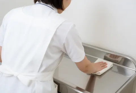 A staff member in a white apron cleans a stainless steel table in a clean clinical setting, focusing on the back and hands.