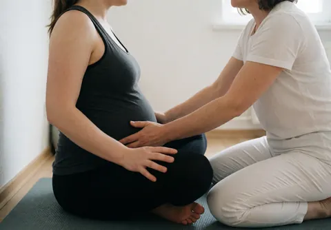 A pregnant woman in black clothing receives a gentle pelvic massage from a professional in white. The scene conveys calm and professionalism with natural light.