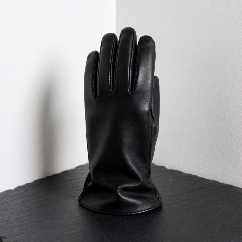 A high-quality black leather glove resting on a dark slate surface with a white wall background and soft lighting highlighting the texture.