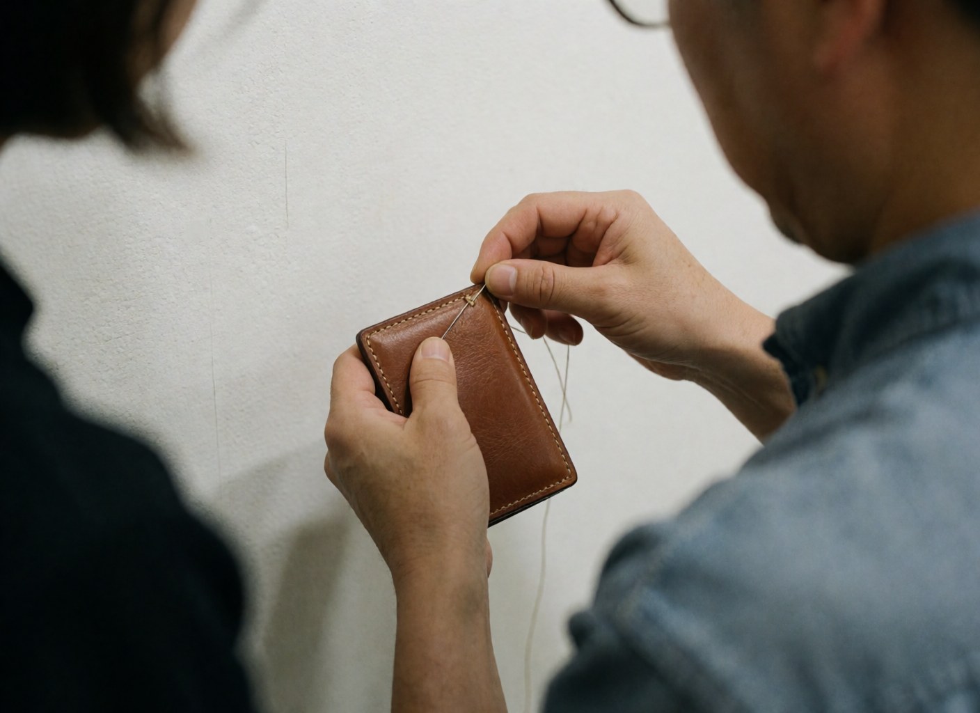 Close-up of an artisan hand-stitching a leather wallet with thread and needle, highlighting the detailed stitching and material texture in a workshop with a white wall.