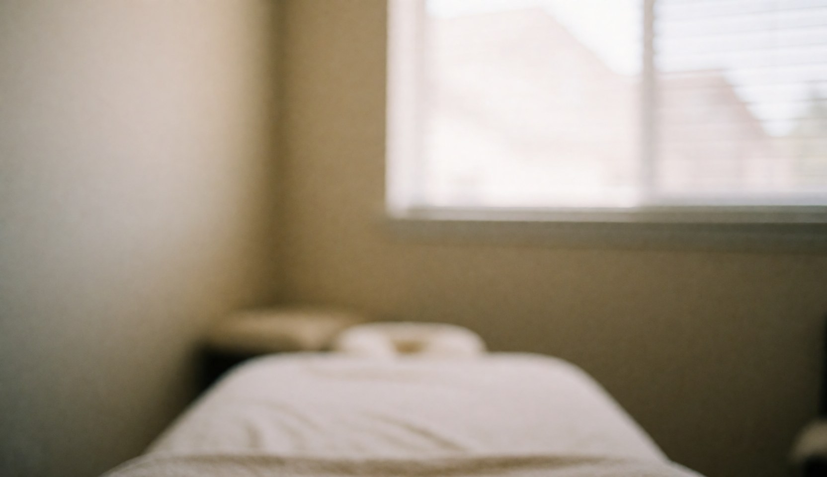 Serene massage room background featuring a white treatment bed, beige walls, and a window with blinds, ideal for text overlays and web design.