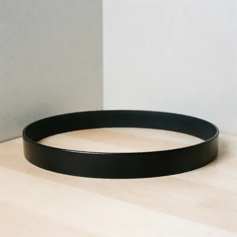 A glossy black leather belt with smooth edges displayed on a light wooden surface against a gray wall with professional lighting.