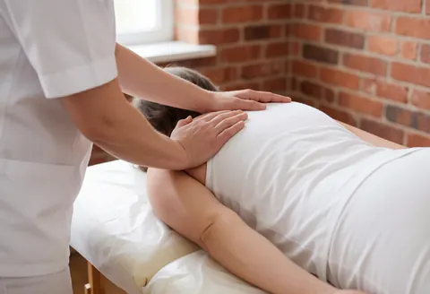 A therapist wearing a white uniform kneads the shoulder area of a client lying on a treatment table with natural lighting