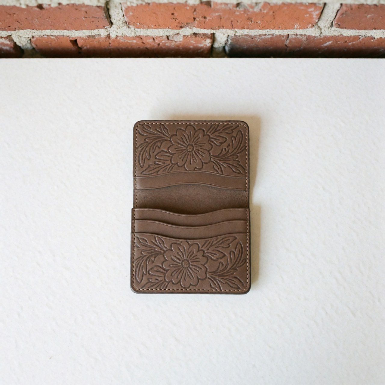 Top view of a brown leather wallet with floral embroidery on a white and red brick background, showcasing clean presentation and visible texture.