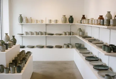An empty ceramic showroom featuring minimalist decor, concrete floors, and natural lighting highlighting various pottery pieces on display shelves.