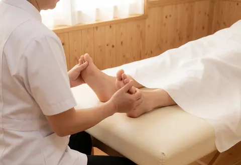 A therapist in a white uniform applies a foot massage to a client resting on a bed with white sheets in a room with wooden walls and natural light.