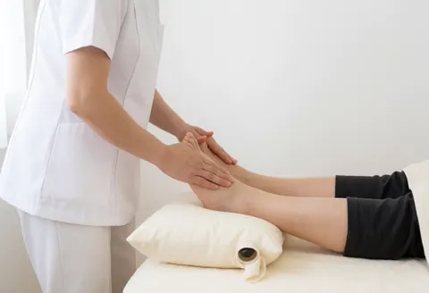 Reflexologist in white uniform performs foot massage on client resting on white pillow in clean professional spa setting