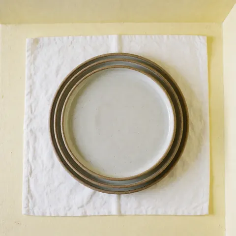 Stacked handmade ceramic plates on white linen cloth against a light yellow wall with natural lighting