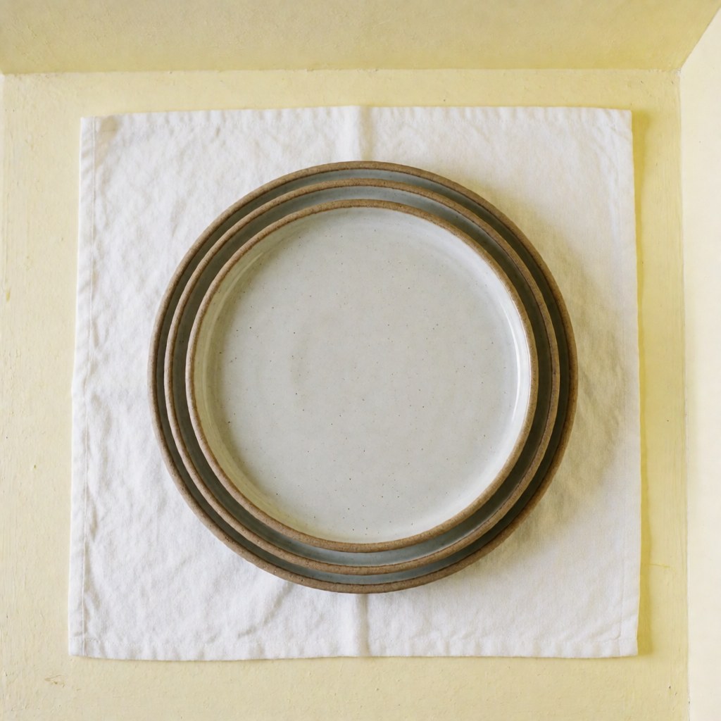 Stacked handmade ceramic plates on white linen cloth against a light yellow wall with natural lighting