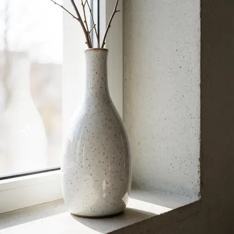 Speckled glazed ceramic vase holding dried twigs on a concrete surface near a window with natural light