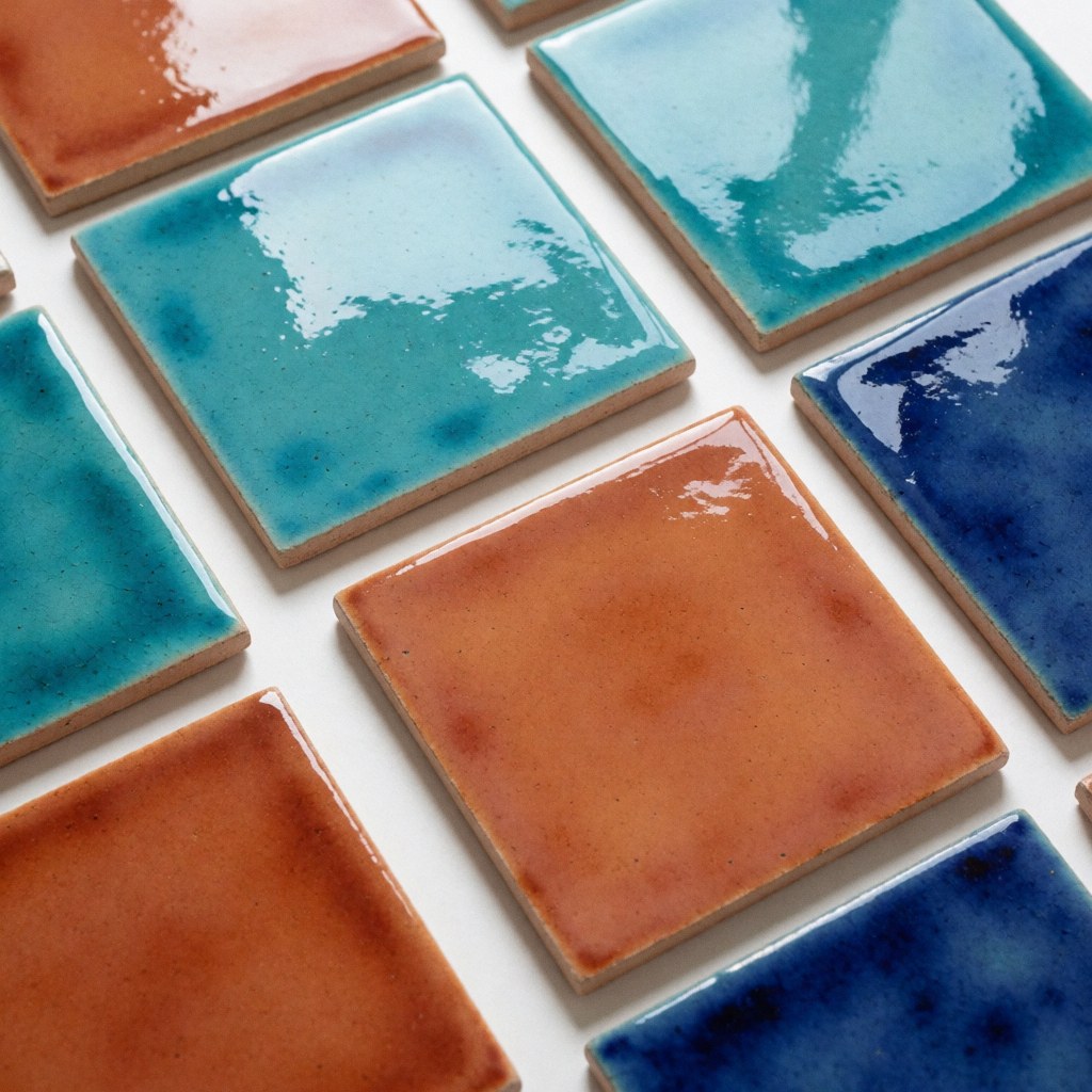 Close-up view of colorful handmade ceramic tiles in turquoise and terracotta shades arranged on a clean white background with natural wood accents