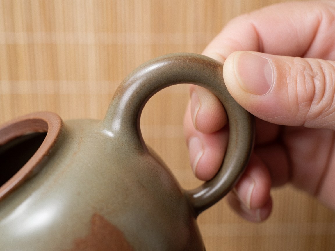 Close-up of a hand holding the handle of a handmade ceramic teapot with earth glaze on a wooden background
