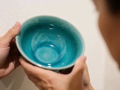Close-up of handmade turquoise glazed ceramic bowl held in hands against a warm wall background, ideal for kitchen and decor.