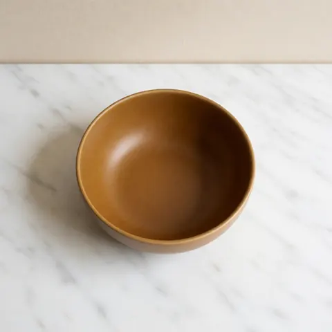 Handcrafted ocre ceramic bowl placed on a white marble surface with a beige wall and soft lighting, suitable for a modern dining table setting.