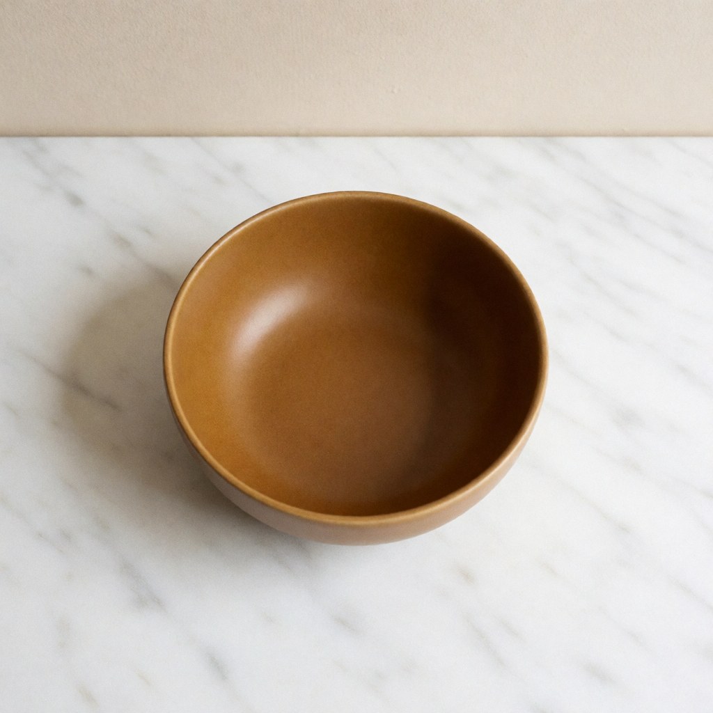 Handcrafted ocre ceramic bowl placed on a white marble surface with a beige wall and soft lighting, suitable for a modern dining table setting.