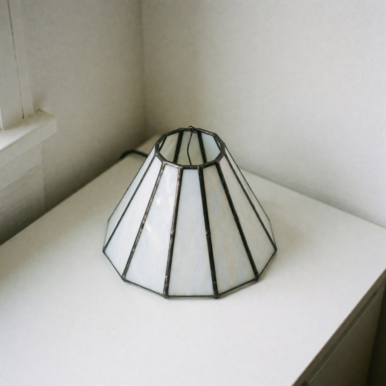 A minimalist white decorative glass lamp with a metal frame placed in a corner against a white wall, emitting soft light for interior design.