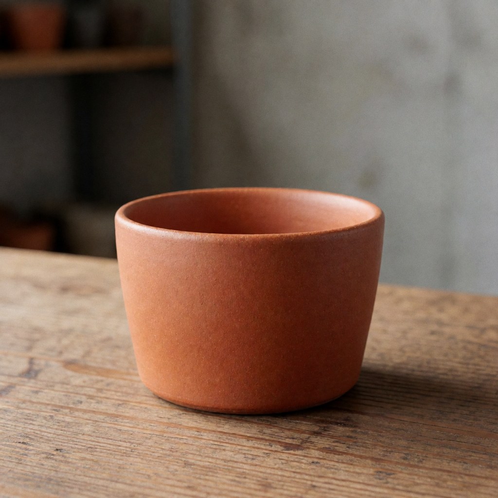 Close-up of a handmade terracotta ceramic coffee mug placed on a rustic wooden table, showcasing natural textures and earthy tones suitable for product photography.