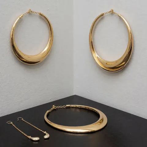 Glossy gold jewelry set featuring a necklace and matching earrings displayed on a black surface against gray walls, showcasing modern elegant design for jewelry stores