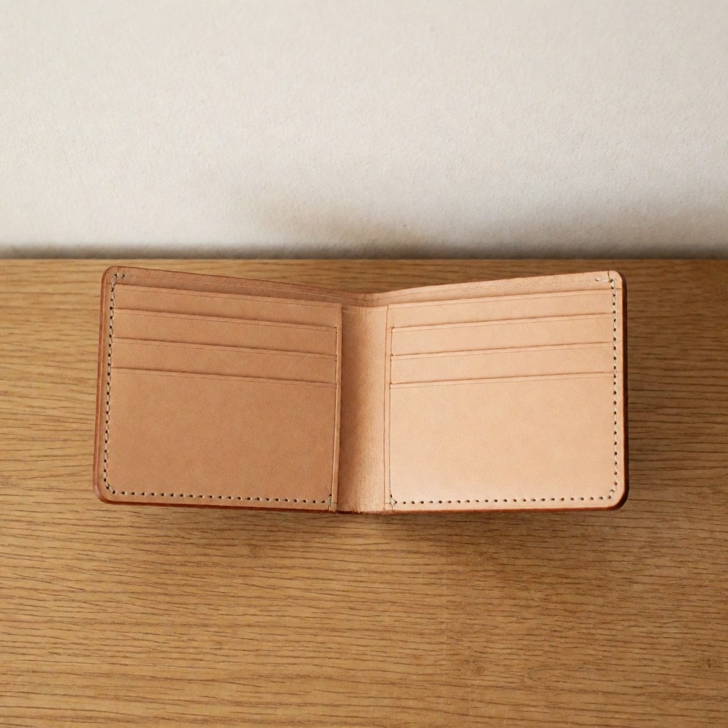 An open brown leather wallet displaying its interior organization and stitching details, resting on a light wooden background